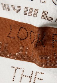 Gestrickter Stoff mit einem Farbgestaltung in Weiß und Braun, verziert mit glänzenden Strasssteinen, die Schriftzüge und Muster bilden. Weiche Textur und ausgeprägte Details.