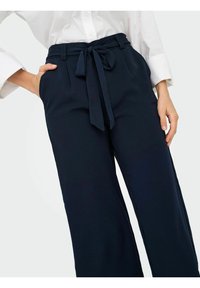 Pantalon larges bleu marine avec une finition texturée, doté d'une taille haute et d'un détail de nœud à l'avant, associé à une chemise blanche boutonnée.