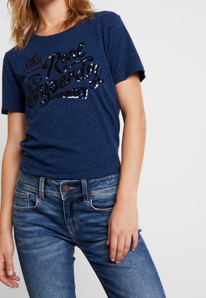 Kvinde iført en blå t-shirt med korte ærmer med sort paliettekst og tætsiddende blå jeans mod en ensfarvet lys baggrund.