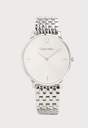Vékony ezüst Calvin Klein karóra minimalista fehér számlappal, ezüst órajelölőkkel és láncos fém szíjjal.