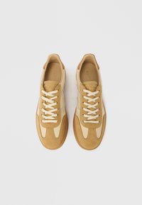 Tan suede and leather sneakers with cream accents, rubber sole, and beige laces. Features a rounded toe and stitched detailing.