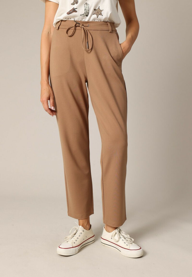 Pantalons marron clair avec une taille à cordon, poches latérales et une coupe fuselée, associés à des baskets blanches. Texture de tissu lisse.