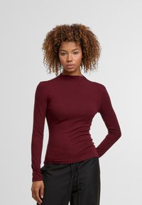 Burgundská halenka s dlouhým rukávem a přiléhavým střihem, vysokým límcem a hladkým povrchem. Kombinována s černými kalhotami, na světlém pozadí.