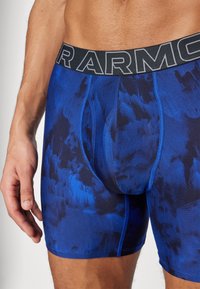 Boxer-briefs bleus à motif avec un design abstrait bleu foncé. Comprend une ceinture noire épaisse avec "ARMOUR" en lettres grises.