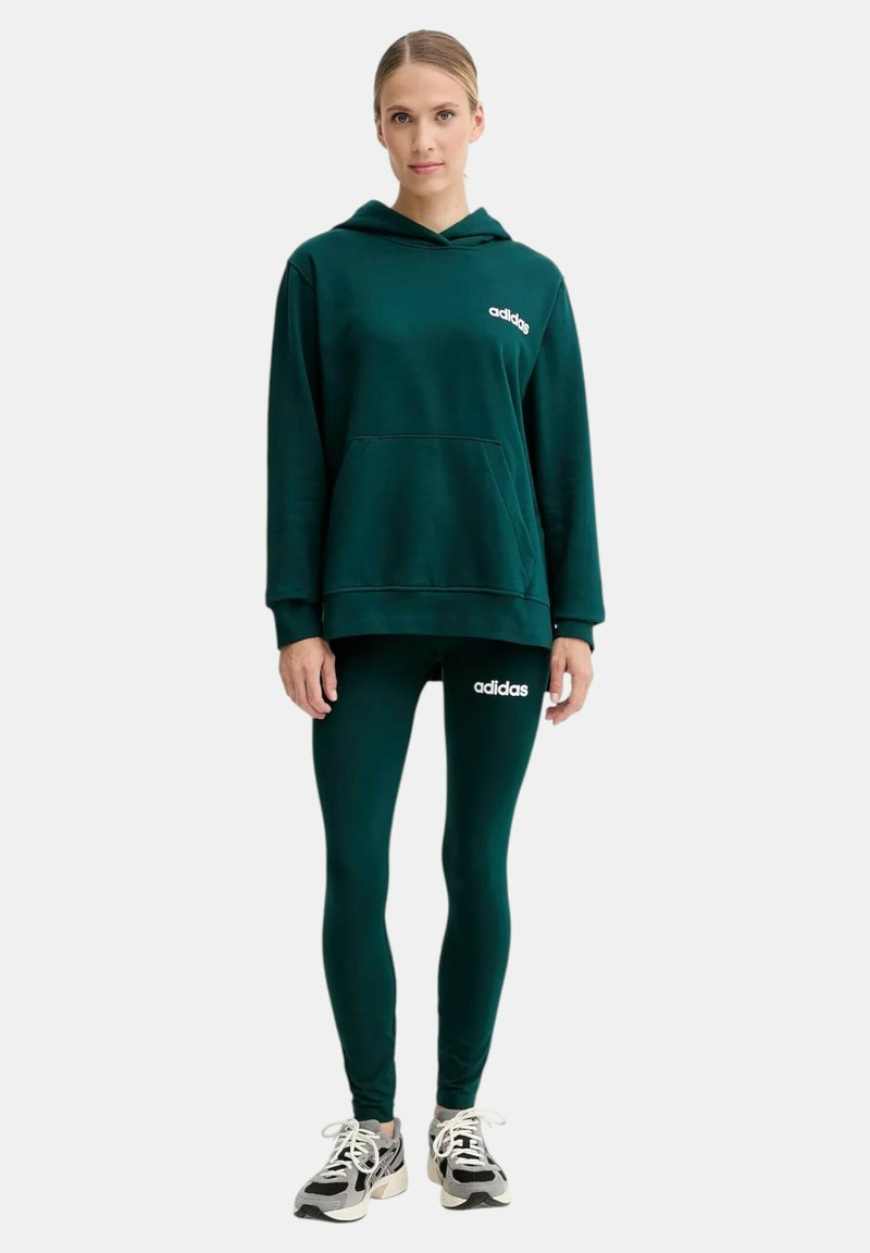 Donna in piedi che indossa un felpa Adidas verde scuro e leggings con logo, abbinati a sneakers bianche e nere, su sfondo bianco uniforme.