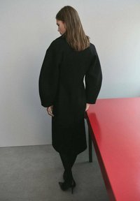 Femme debout à l'intérieur près d'une table rouge, vêtue d'un long manteau noir et de bottes noires à talons, faisant face à un mur gris.