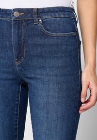 Tumed sinine denimist pükste slim fit stiil. Omadusteks on esikott, metallnupp ja kontrastsed õmblused õmbluste ääres. Sile tekstuur.