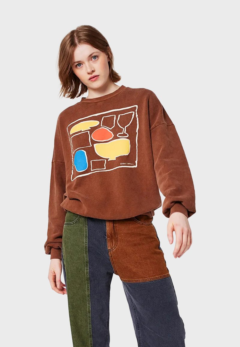 Joven vistiendo sweatshirt marrón con formas abstractas coloridas y pantalones de patchwork en verde, azul y marrón, contra un fondo liso.