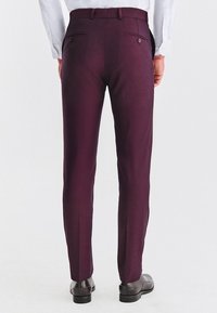 Mężczyzna ubrany w dopasowane, ciemnofioletowe spodnie, białą koszulę wpuszczoną w spodnie oraz ciemnobrązowe eleganckie buty, stojący tyłem do widza.