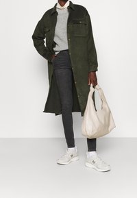 Abrigo largo verde oliva con botones, suéter gris, jeans oscuros, zapatillas blancas con acentos grises y un bolso suave de color beige claro.