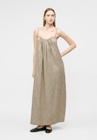 Ženska s ravnimi rjavimi lasmi nosi brezrokavno bež maxi obleko z gubami in črne balerinke, stoji z roko na boku.