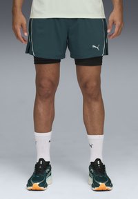 Puma sportshorts in donkergroen, met witte accenten, gecombineerd met zwarte compressieshorts, witte sokken en hardloopschoenen.