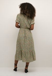 Zelena cvetlična obleka s kratkimi, napihnjenimi rokavi, elastičnim pasom in plastnatim robom s pompom. Vsebuje rožnate in rumene cvetlične vzorce.