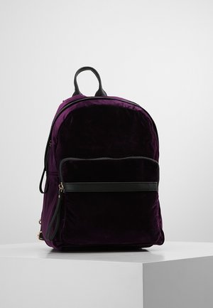 Lilla fløjlsrygsæk med sort læderhåndtag, lynlås på frontlommen og sidestrimmel, placeret på en hvid piedestal mod en ensfarvet baggrund.