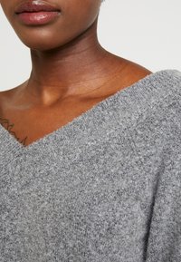 Pull en maille gris avec un large col en V. Texture douce visible avec une finition légèrement duveteuse. Gros plan sur l'encolure et l'épaule.