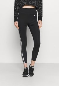 Leggings neri con strisce laterali bianche, vita alta e tessuto elasticizzato. Abbinati a scarpe sportive nere e a un crop top nero stampato.