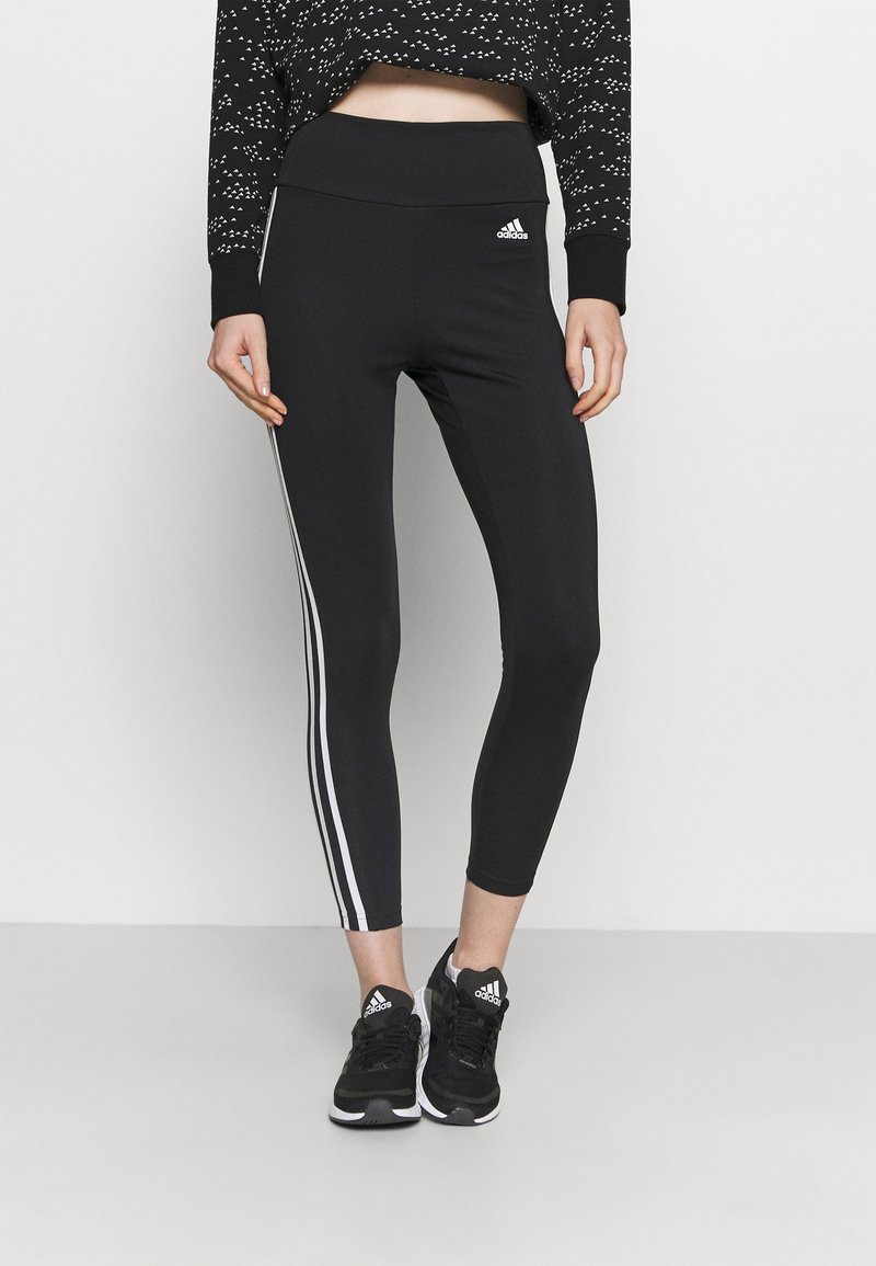 Leggings neri con strisce laterali bianche, vita alta e tessuto elasticizzato. Abbinati a scarpe sportive nere e a un crop top nero stampato.