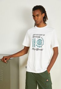 Witte katoen t-shirt met afgeprinte tekst in groen en zwart. In combinatie met olijfgroene cargo-broeken en een metalen locker op de achtergrond.