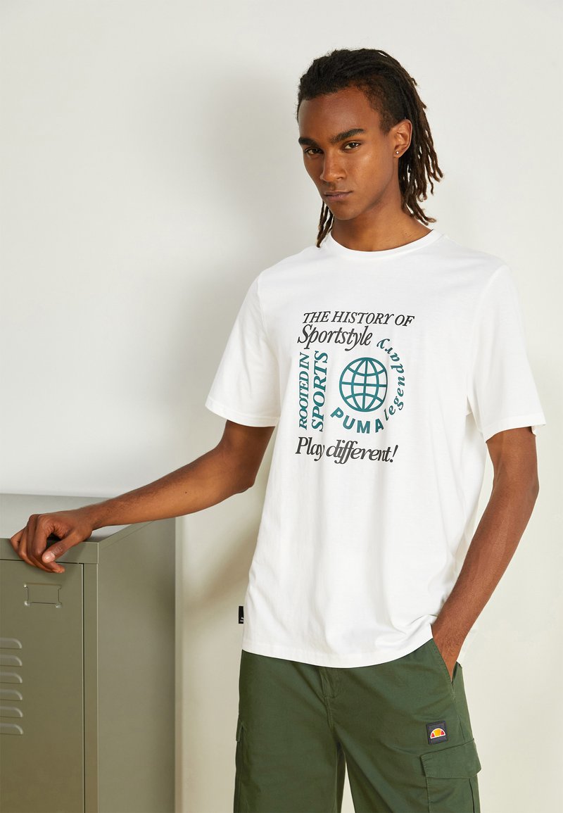 Witte katoen t-shirt met afgeprinte tekst in groen en zwart. In combinatie met olijfgroene cargo-broeken en een metalen locker op de achtergrond.