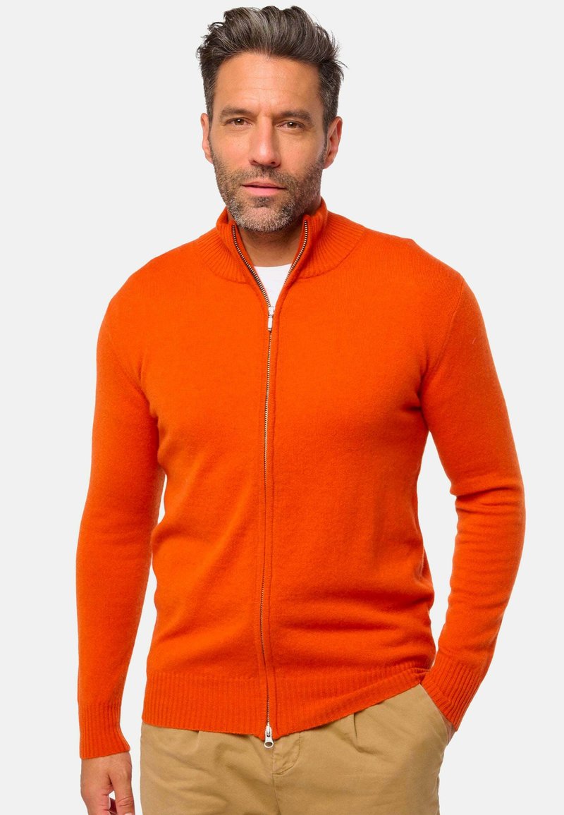 Un pull zippé orange avec un col montant, des poignets et un ourlet côtelés, fabriqué à partir d'un tissu texturé, porté sur une chemise blanche.