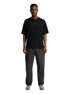 Mężczyzna stoi twarzą do przodu, ubrany w luźną czarną koszulkę z numerem "223" na piersi, ciemnoszare spodnie i jasnoszare trampki na białym tle.