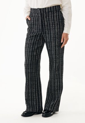 Person trägt schwarz-weiß karierte weite Hosen, weißes Hemd und schwarze glänzende Loafers, steht mit einer Hand in der Tasche.