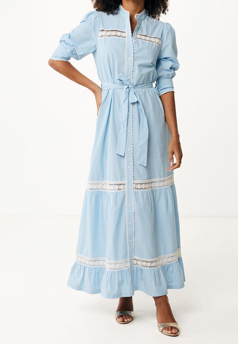 Femme portant une longue robe bleu clair ceinturée, avec des empiècements en dentelle, des manches bouffantes et des chaussures argentées à bout ouvert, debout devant un fond uni.