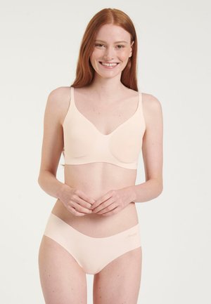 Ung kvinde med rødt hår, der smiler, iført lysebeige sømløs bh og matchende undertøj, stående mod en ensfarvet lys baggrund.