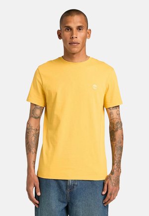 Mann mit kurzen Haaren, der ein gelbes kurzärmeliges T-Shirt und blaue Jeans trägt, steht mit entspannt an den Seiten hängenden Armen vor einem einfarbigen Hintergrund.