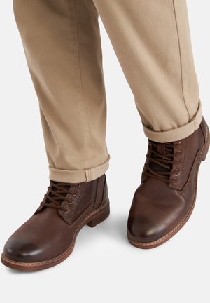 Botas marrones de cuero con cordones, usadas con pantalones beige remangados, sobre un fondo blanco, mostrando vistas frontal y lateral del calzado.