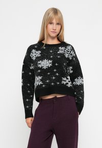 Schwarzer, cropped Pullover mit Schneeflockenmuster in Silber. Verfügt über einen gerippten Halsausschnitt und Bündchen. Kombiniert mit dunkelvioletten, hoch taillierten Hosen.