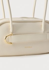 Witte leren handtas met textuur, voorzien van een gouden accent, dubbele handgrepen en geperst merklogo aan de zijkant. Minimalistisch ontwerp.