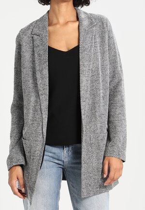 Frau trägt ein graues Blazer mit Fischgrätmuster über einem schwarzen Oberteil und hellblauen Jeans, steht mit entspannten Händen an den Seiten.