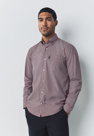 Mees, kes kannab punase-valge ruudulise nööbitava särgi ja tumedaid pükse, seisab käed taskutes tavalise tausta ees.