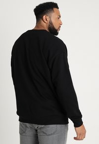 Sudadera negra de cuello redondo con puños y dobladillo acanalados, hecha de una tela suave. Modelo usando jeans grises, visto desde la espalda.