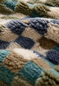 Mjukt fleecetyg med ett texturerat, mångfärgat lapptäcksmönster i blå, beige, bruna och gröna toner.