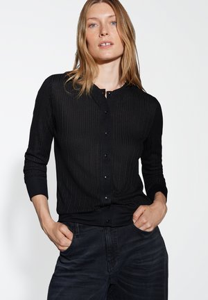 Schwarzer gerippter Cardigan mit rundem Halsausschnitt, langen Ärmeln und Knopfverschluss. Weicher Stoff und taillierte Passform, kombiniert mit dunklen Jeans.