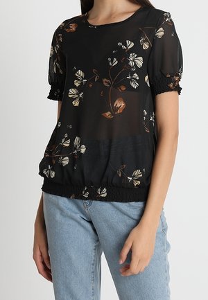 Schwarze transparente Bluse mit weißem und braunem Blumenmuster, kurzen Puffärmeln mit elastischen Bündchen und gesammeltem elastischem Saum, getragen zu hellblauen Jeans.