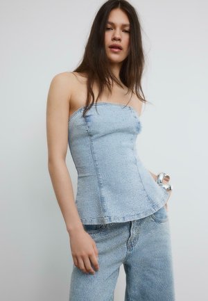 Kvinde iført lyseblå strapless denimtop og matchende jeans med små rhinsten, stående mod en enkel lys baggrund.