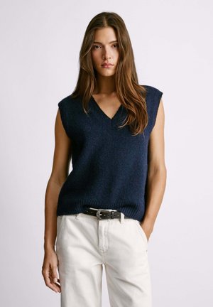 Maglione senza maniche in maglia blu navy con scollatura a V, realizzato in filato morbido, abbinato a pantaloni a vita alta di un bianco sporco e una cintura con dettagli in argento.