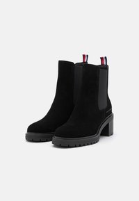 Botines de ante negro con paneles laterales elásticos, un tacón grueso y una suela con relieve. Presenta una lengüeta en la parte posterior en colores rojo, blanco y azul.