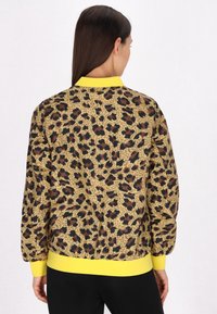 Leopardprint bomberjack in doorschijnend materiaal, met een gele ribgebreide kraag en manchetten. De achterkant toont het patroon met bruin en zwarte accenten.