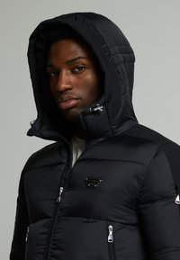 Schwarze Pufferjacke mit einer Kapuze zum Reißverschluss, glänzenden Reißverschlüssen, einem Logopatch und einem gesteppten Textur. Der Körper ist isoliert und figurbetont.