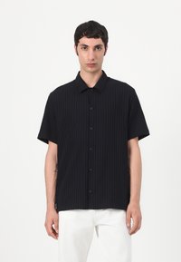 Camicetta nera a maniche corte con bottoni, caratterizzata da un motivo a coste testurizzate e colletto classico. Indossata con pantaloni bianchi.