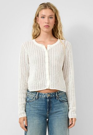 Vrouw met lang blond haar draagt een witte, gebreide cardigan met knopen en een blauw spijkerbroek met hoge taille tegen een effen achtergrond.