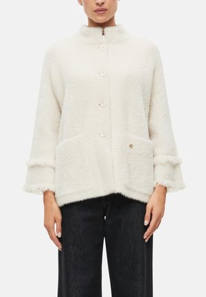 Vrouw draagt een witte, donzige jas met knopen, wijde mouwen en voorzakken, gecombineerd met een zwarte broek, tegen een witte achtergrond.
