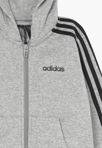 Grijze rits-hoodie gemaakt van zachte stof, met drie zwarte strepen langs de mouwen en een geborduurd zwart logo op de borst.