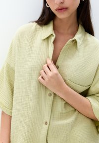Lichtgroen, textuurshirt met een knoopsluiting aan de voorkant en korte mouwen. Het heeft een zak en een ontspannen pasvorm.