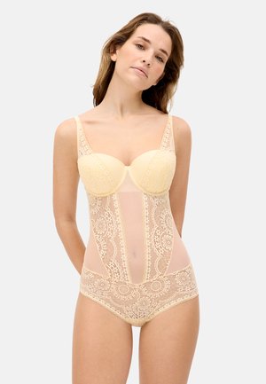 Mujer con body de encaje color beige con copas con aro, patrones florales y paneles de malla, posando frente a un fondo claro y liso.