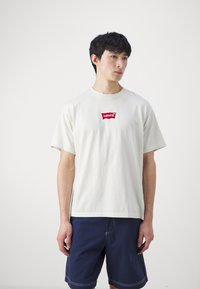Trumpas rankovėmis, kreminės spalvos marškinėliai iš medvilnės su raudonu Levi's logotipu ant krūtinės, derinami su tamsiai mėlynomis šortais, turinčiais kontrastuojančius siūlus.